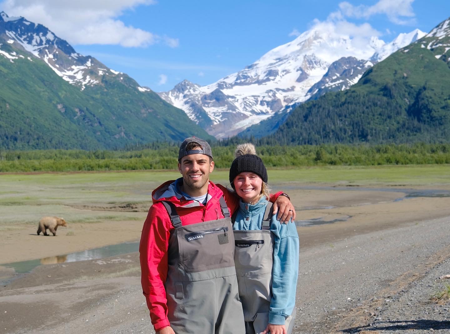 Tessa Wisniewski鈥�17, 鈥�19 and her husband standing on a shoreline with a bear and mountains behind them